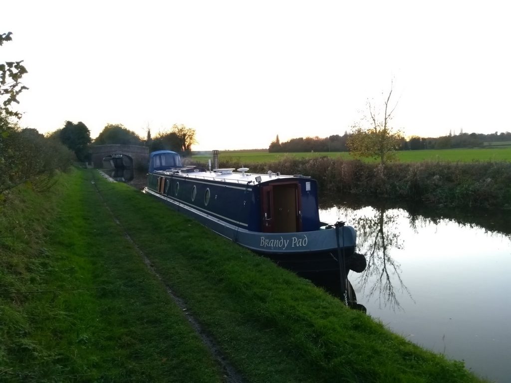 A photo of Brandy Pad moored against a green bank and sun setting behind. Smoke from chimney is visible. Cosy