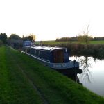 A photo of Brandy Pad moored against a green bank and sun setting behind. Smoke from chimney is visible. Cosy